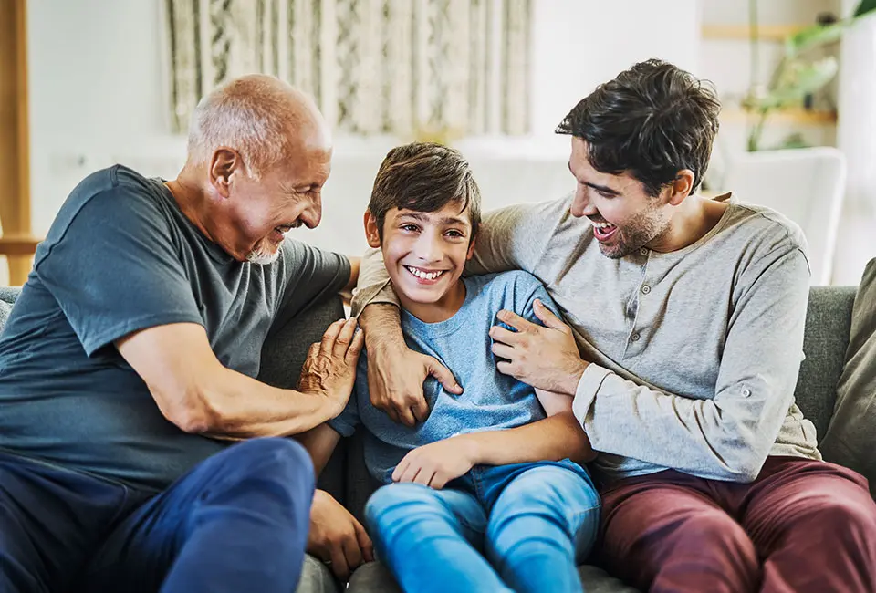 A boy sits on a couch with his family, all smiling in a relaxed living room setting.