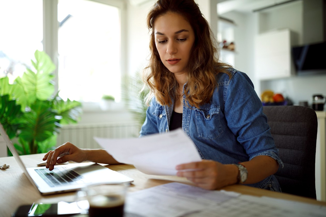 Young woman sorting her finances across paperwork and laptop at home.jpg